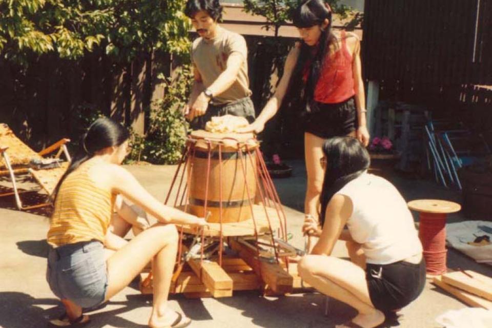 Two people crouched on the ground and two people standing preparing to stretch a drum head on the drum in the middle of them